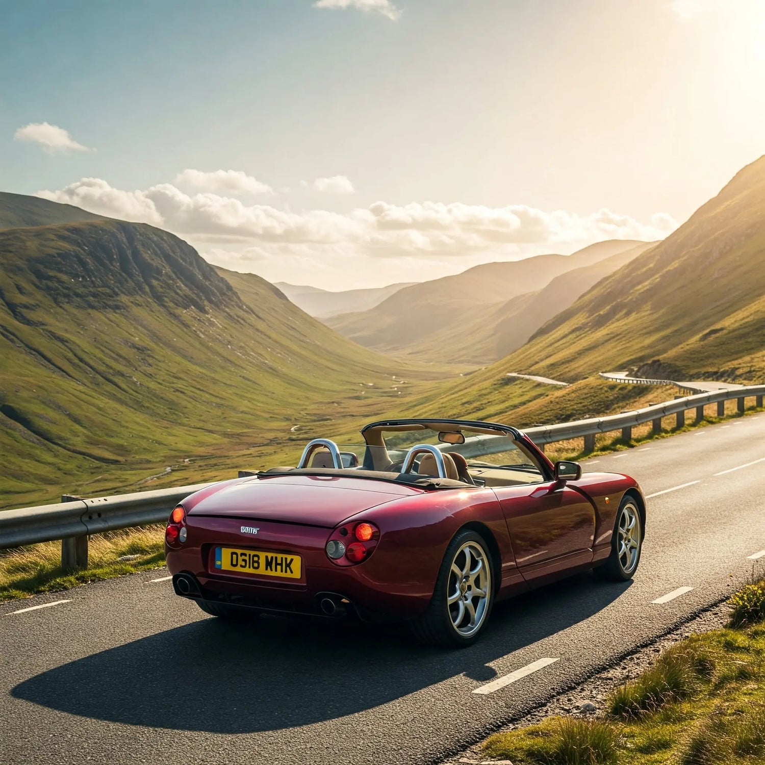 A classic burgundy TVR Chimaera convertible driving along a winding mountain pass road under a bright, hazy sun.